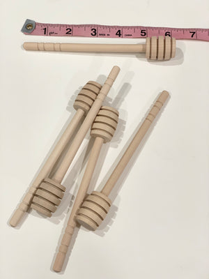 Natural unfinished wooden honey dippers arranged on white surface with pink measuring tape above for size reference