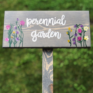 Large custom wooden garden sign painted gray with white cursive "perennial garden" letters and colorful floral illustrations on wood grain background