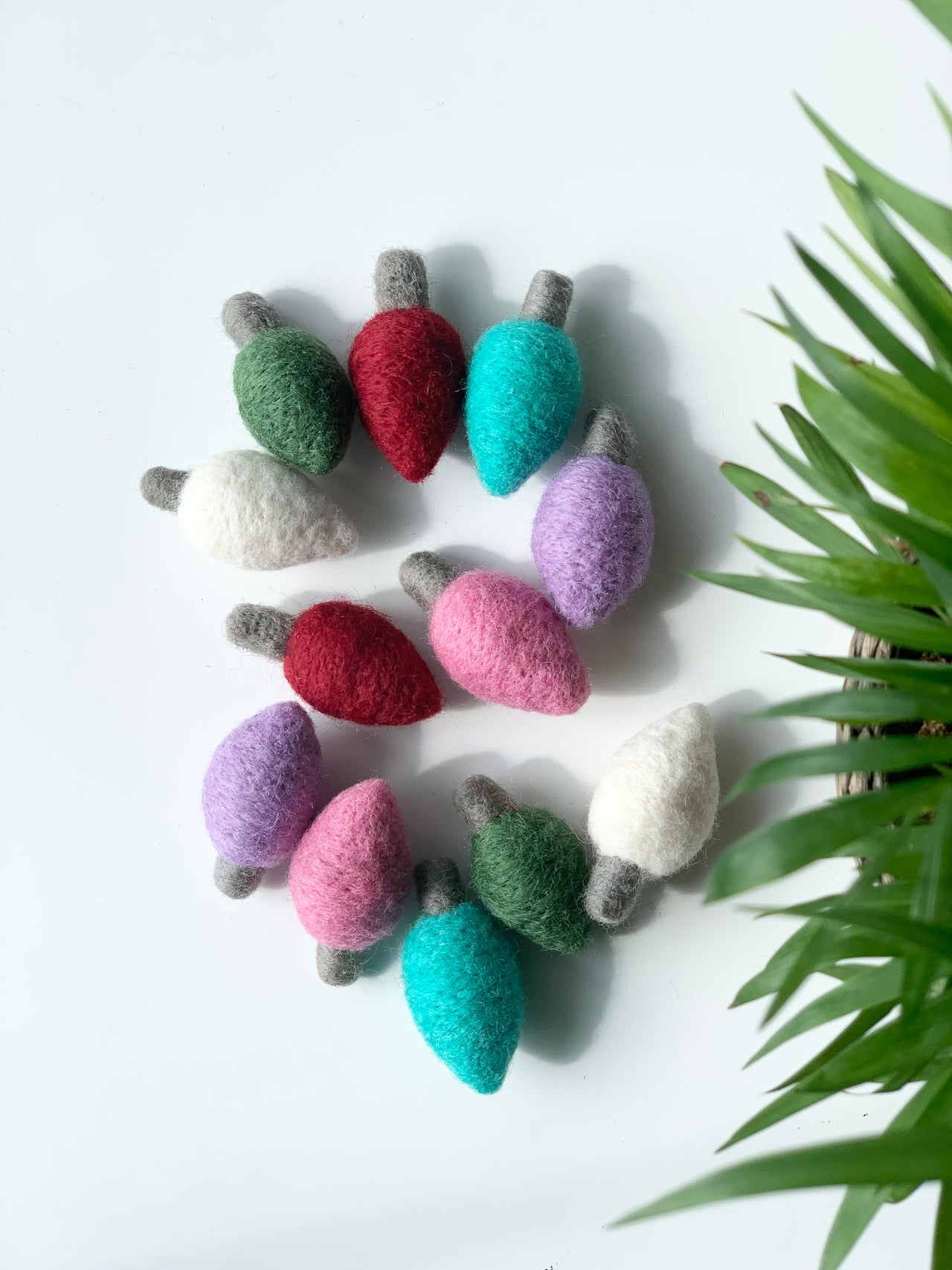 Collection of colorful handmade felt light bulbs arranged in circle on white surface with green plant leaves