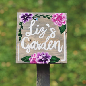 Square wooden garden sign with white cursive letters and hand-painted pink and purple flowers on natural wood background
