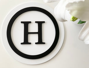 Round 3D monogram sign with black letter H and circle on white two-layer MDF background with white flower accents