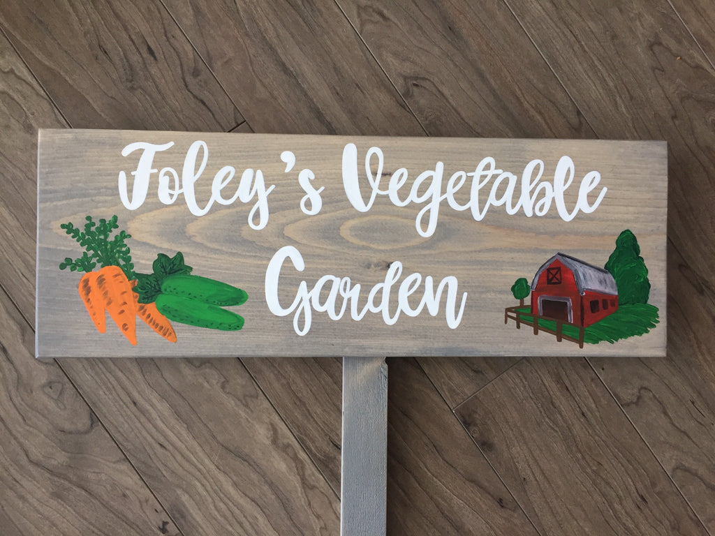 Custom wooden garden sign painted gray with white cursive letters and illustrations of carrots cucumbers and barn on wood grain background