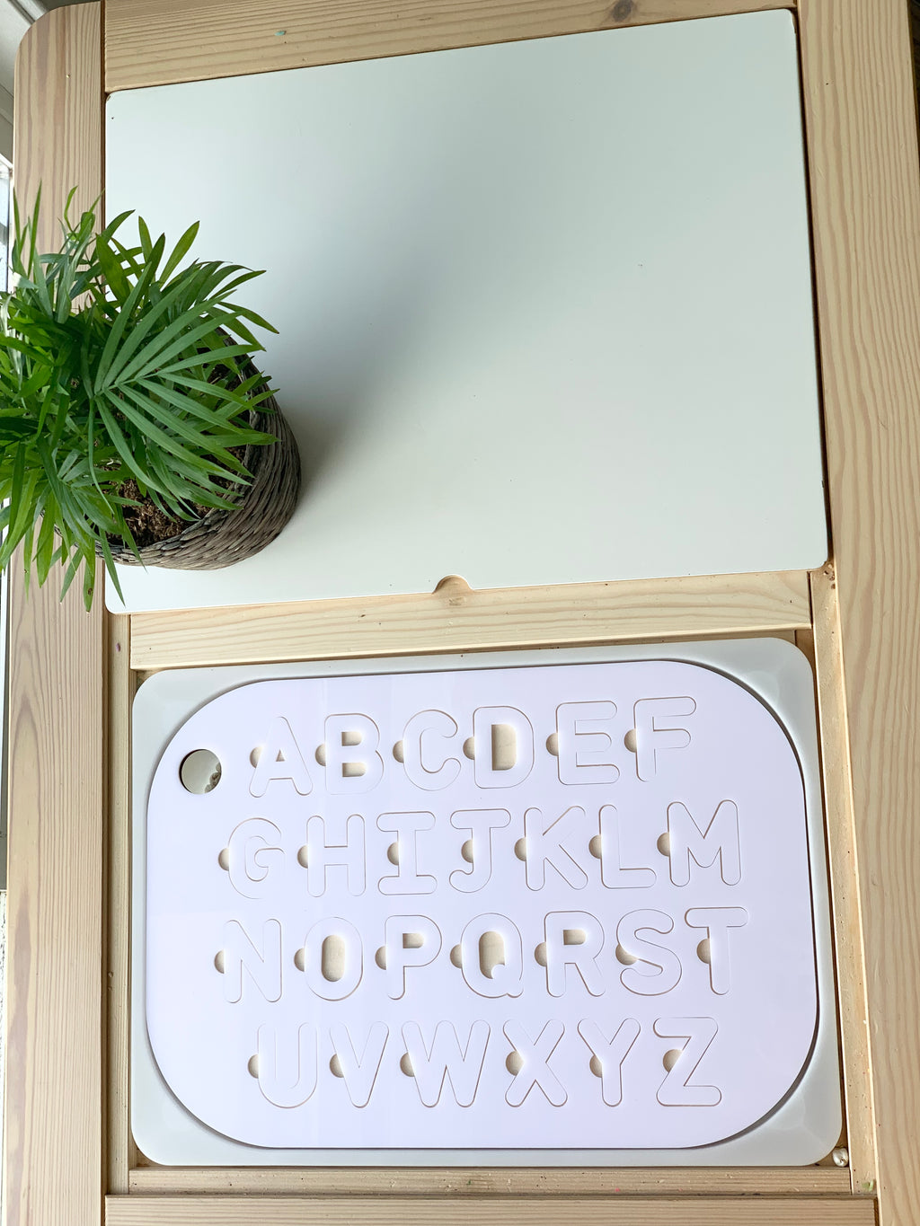 Maple wood Flisat table top insert with white acrylic uppercase alphabet puzzle pieces inside wooden frame with green plant nearby