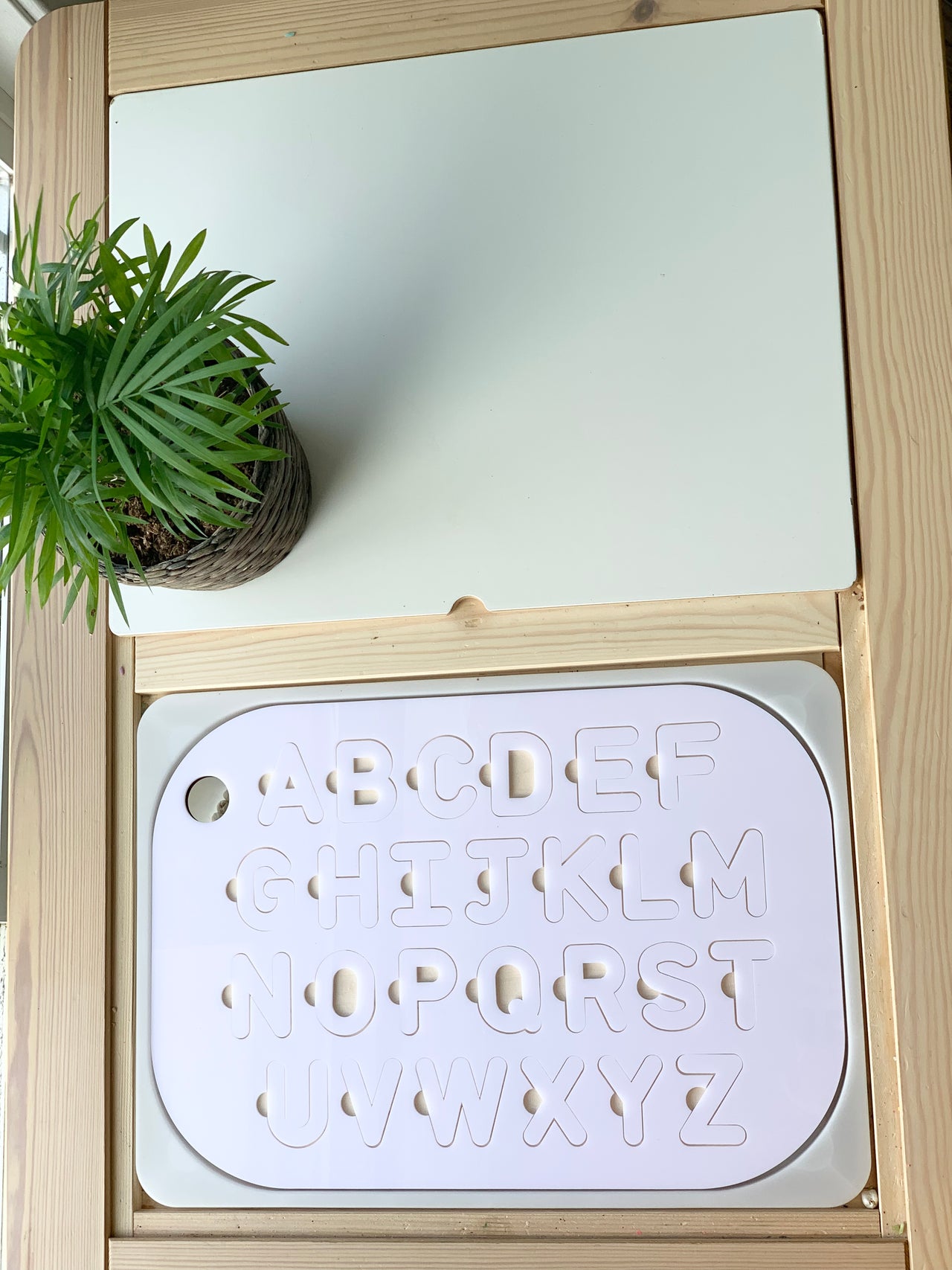 Maple wood Flisat table top insert with white acrylic uppercase alphabet puzzle pieces inside wooden frame with green plant nearby
