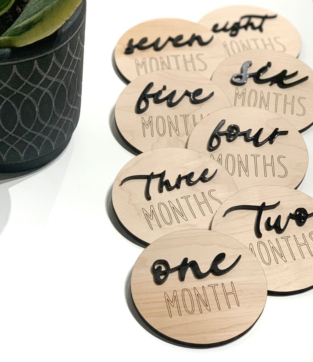 Set of natural wooden round milestone discs with black cursive numbers and engraved MONTHS text on white surface near plant pot