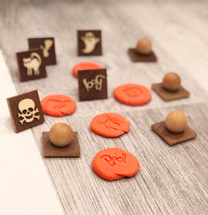Small wooden Halloween play dough stamps with ghost, skull, cat, and "Boo" impressions on orange dough on wood surface