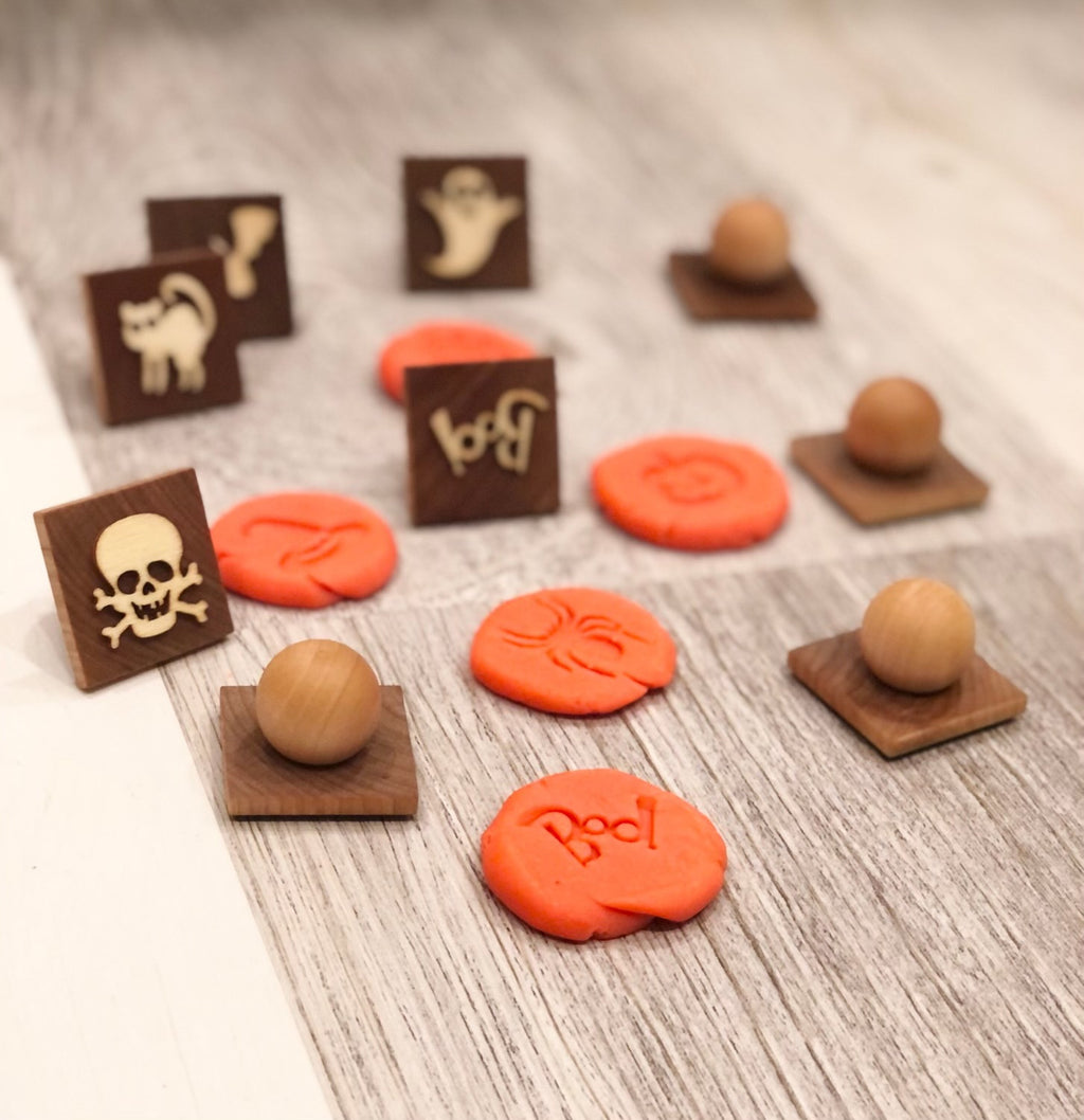 Small wooden Halloween play dough stamps with ghost, skull, cat, and "Boo" impressions on orange dough on wood surface