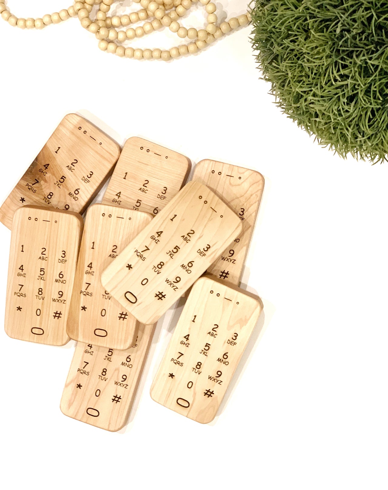 Set of natural wooden toy phones with engraved numbers and symbols on a white surface with greenery and beads nearby