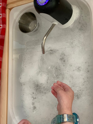 White play kitchen sink filled with bubbles and water pouring from black faucet into clear cup held by child’s hands