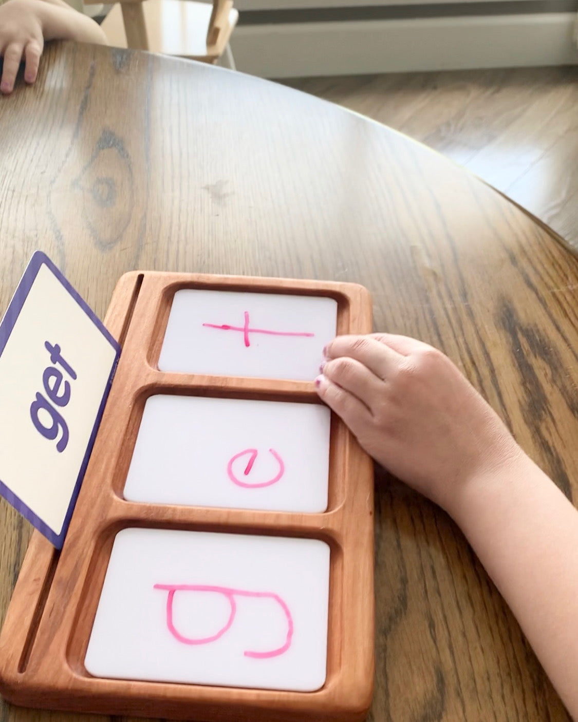 Child's hand using wooden flash card holder with three dry erase cards spelling the word get on wooden table