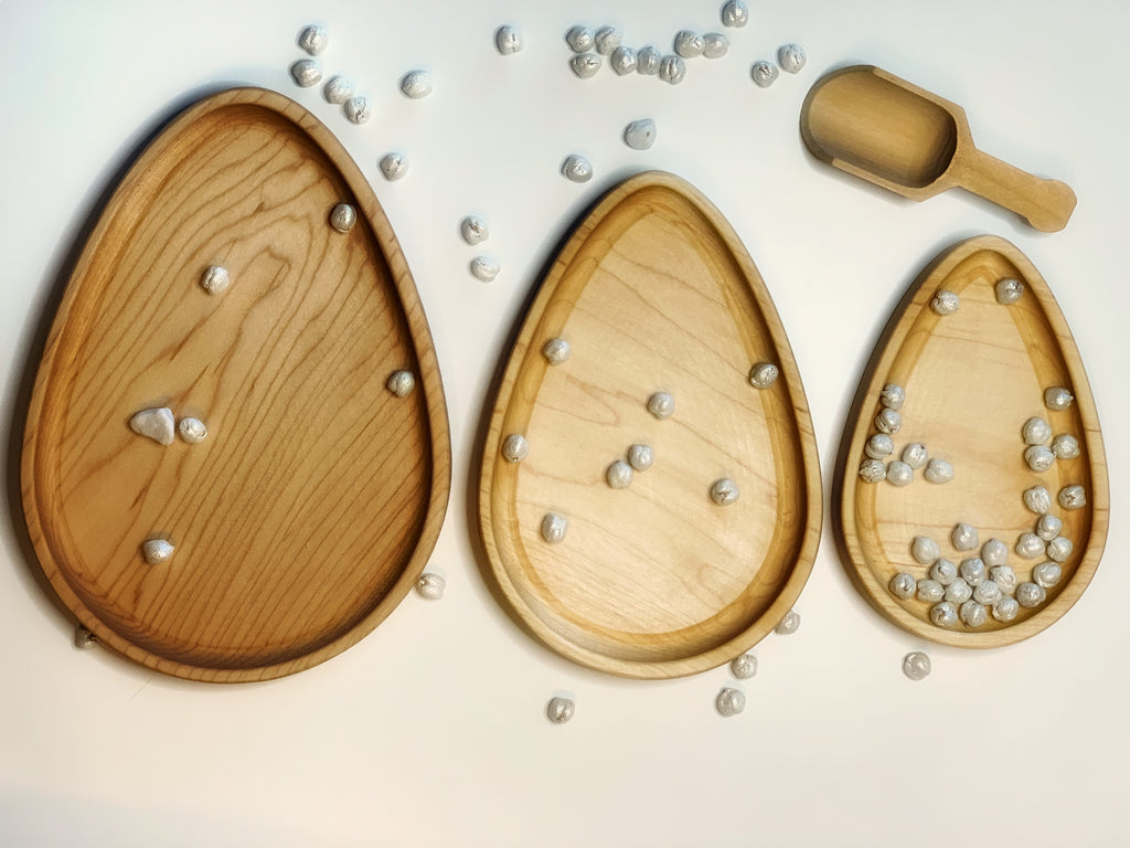 Three wooden nesting egg-shaped trays in varying shades with scattered small white beads and wooden scoop on white background