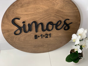 Oval custom wooden sign with black cursive last name and date on natural wood background next to white orchid plant