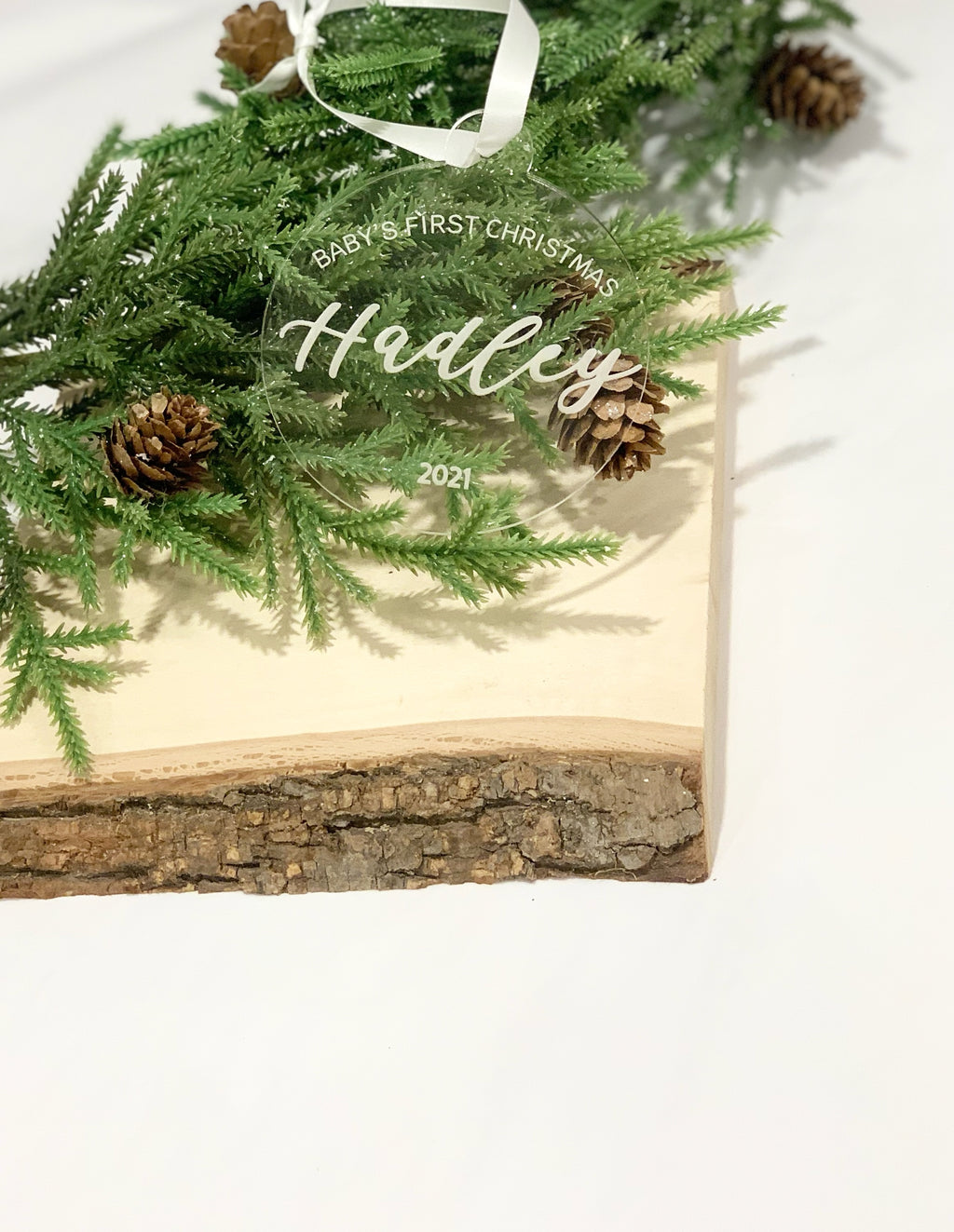 Clear acrylic baby’s first Christmas ornament engraved with name and year, displayed on pine and wood block background