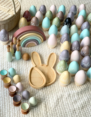 Assorted hollow fillable wooden Easter eggs with pastel colors and cursive names, wooden bunny tray and rainbow stacker on textured surface