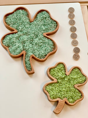 Two wooden shamrock-shaped sensory trays filled with green-dyed rice and wooden letter tiles spelling shamrock on white surface