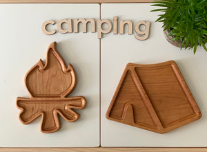 Two wooden sensory trays shaped like a campfire and tent with natural wood grain on white surface with plant and camping letters