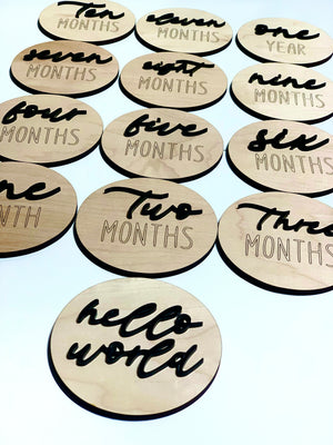 Set of round wooden 3D monthly milestone discs and one "hello world" disc with black cursive and engraved lettering on white background