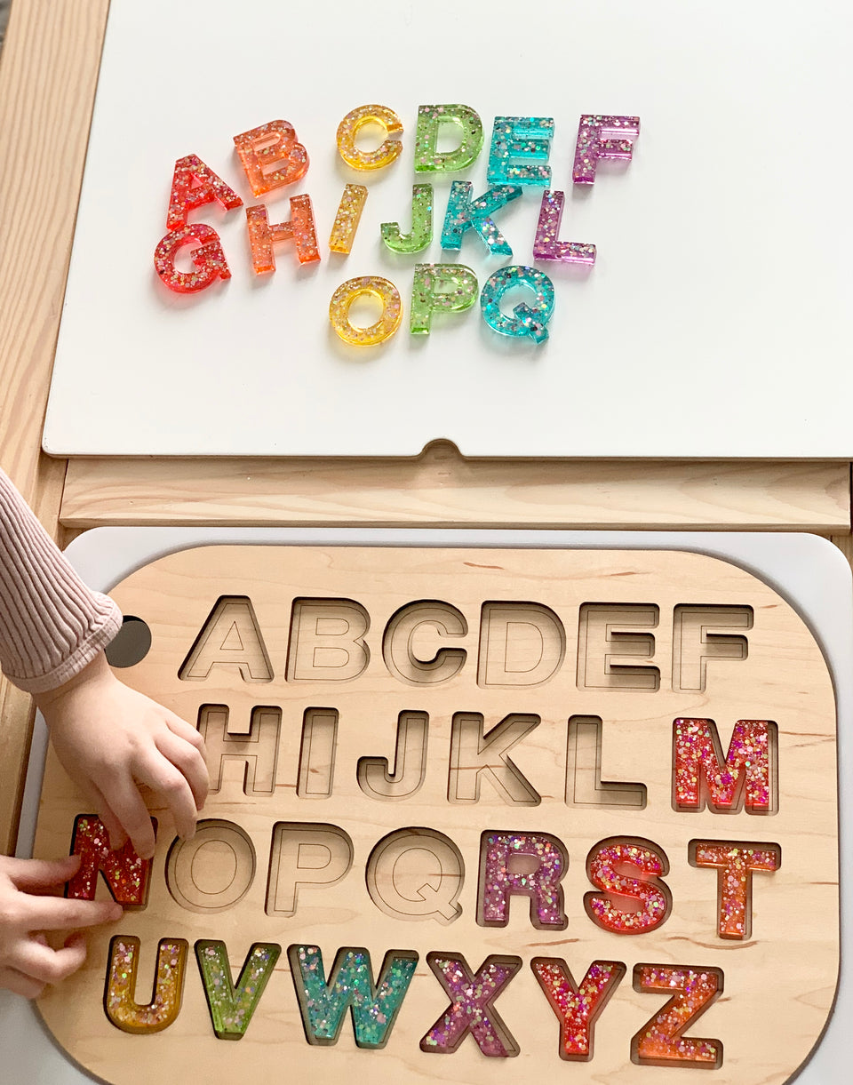 Wooden uppercase alphabet puzzle insert with glittery resin letters arranged on and above the board by child’s hand