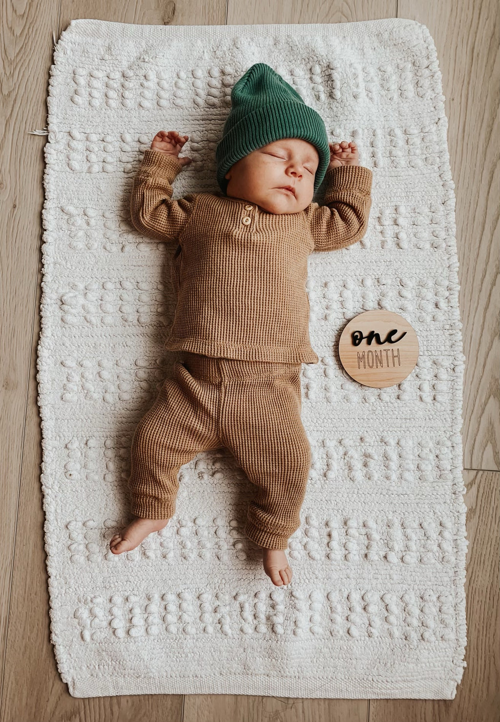 Newborn baby in brown knit outfit and green hat lying on textured white blanket next to one month milestone wooden disc