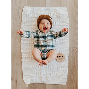Smiling baby wearing brown knit hat and plaid outfit lying on white textured rug with seven months milestone disc on wood floor