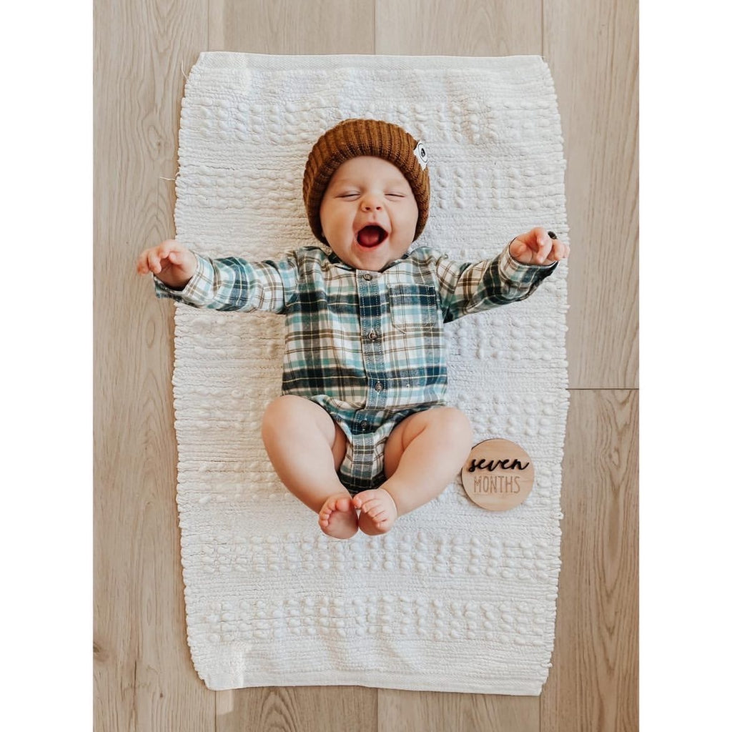 Smiling baby wearing brown knit hat and plaid outfit lying on white textured rug with seven months milestone disc on wood floor