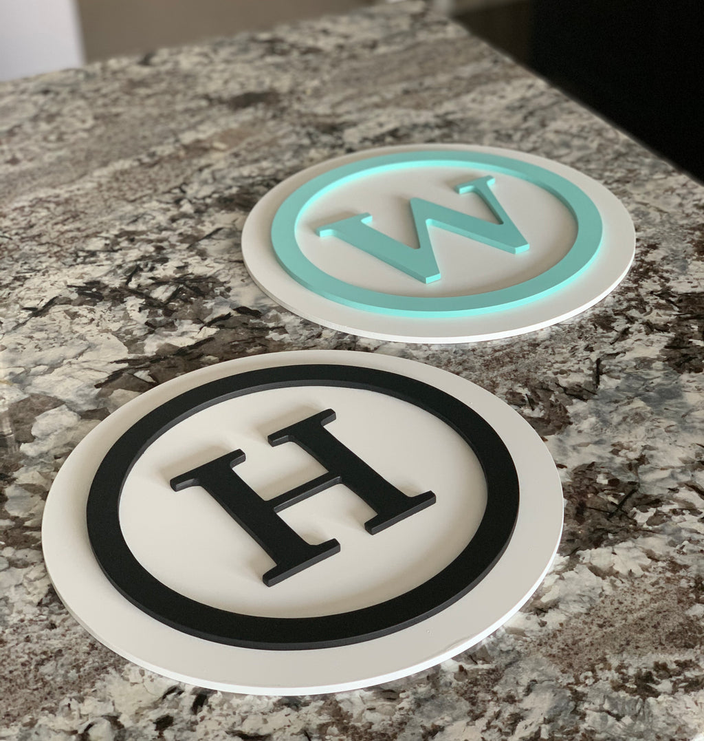 Two 3D monogram round signs made from layered MDF with black and teal letters on white circular backgrounds