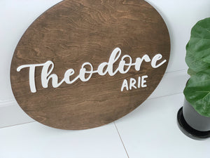 3D round wooden name sign with white cursive and block letters on dark stained wood background near green plant