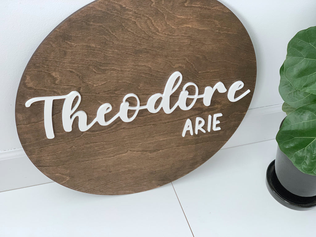 3D round wooden name sign with white cursive and block letters on dark stained wood background near green plant