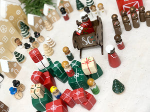 Hand-painted Christmas peg dolls including Santa, Mrs Claus, elves, gifts, trees, and gingerbread houses on white surface