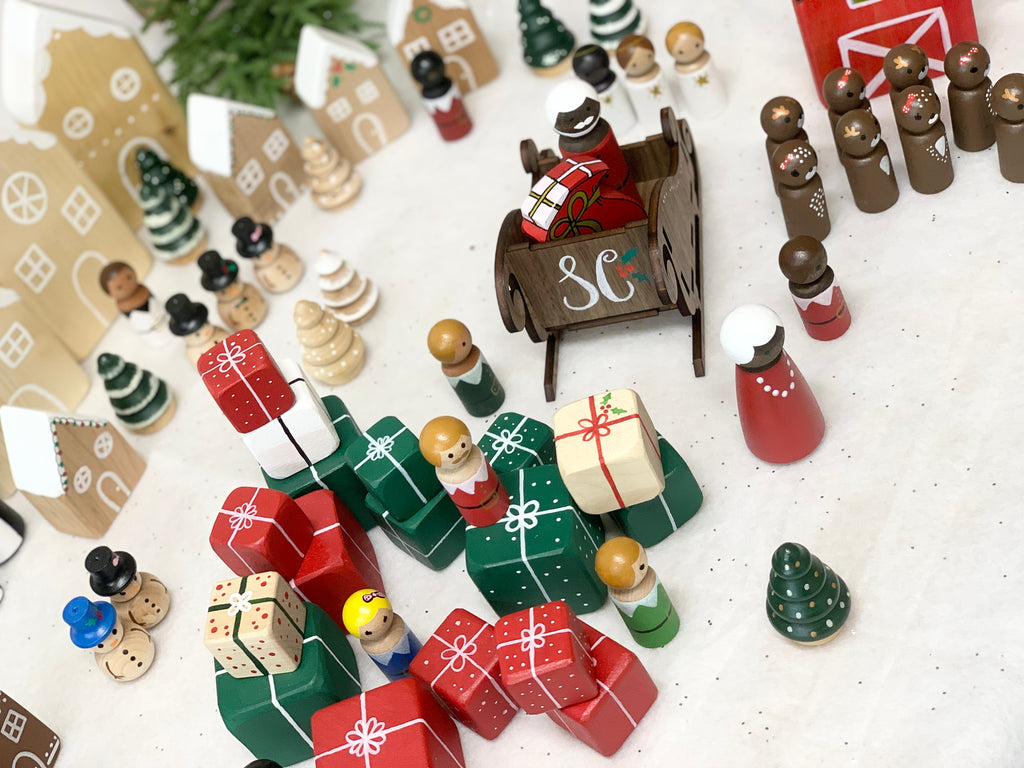 Hand-painted Christmas peg dolls including Santa, Mrs Claus, elves, gifts, trees, and gingerbread houses on white surface