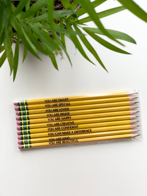 Set of 10 yellow pre-sharpened affirmation pencils with black motivational phrases and pink erasers on white surface near green plant