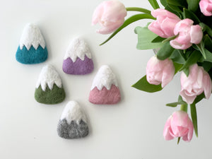 Five handmade felt mountains in blue, purple, green, pink, and gray with white snowy tops on white background with pink tulips