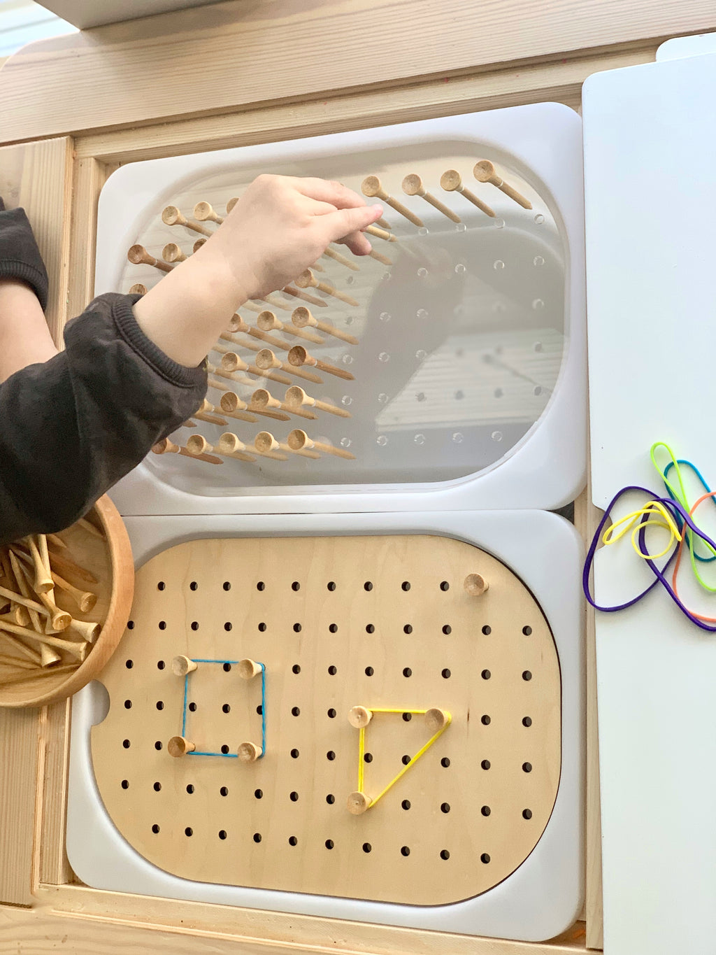 Child’s hand placing wooden golf tees into a Flisat peg board insert with elastic bands and wooden bowl on light wood surface
