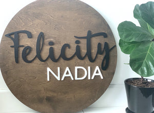 Round wooden 3D name sign with cursive black Felicity and block white Nadia letters on natural wood background