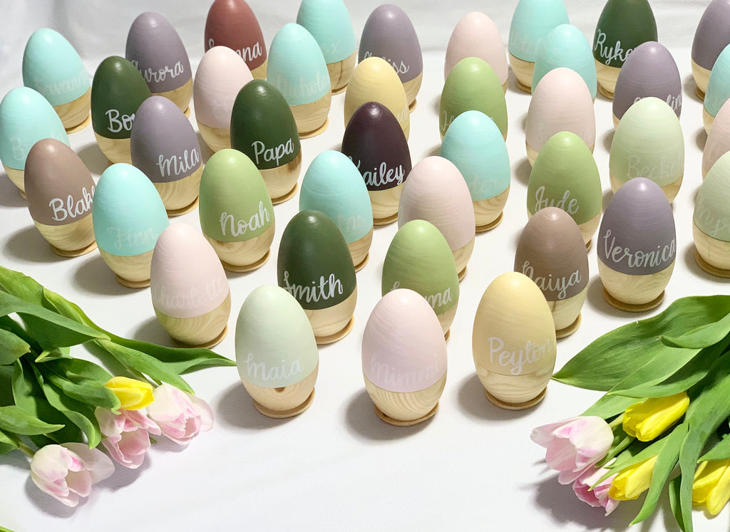Assorted hollow fillable wooden Easter eggs in pastel colors with personalized white script names on natural wood bases