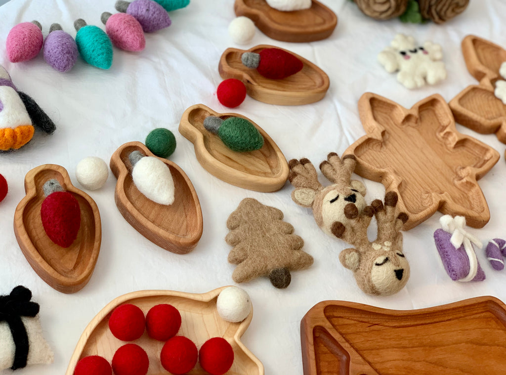 Small natural wooden sensory trays shaped like leaves, tree, and reindeer surrounded by colorful felt balls and figures on white fabric