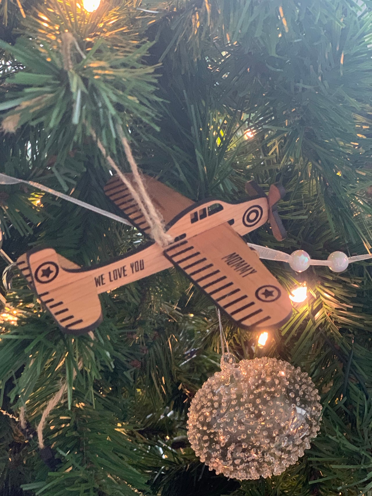 Wooden airplane Christmas ornament with personalized text hanging on decorated green Christmas tree branch