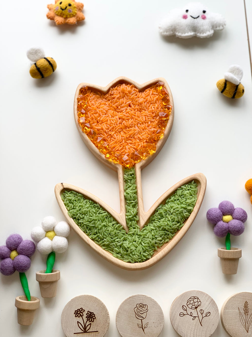 Wooden tulip-shaped sensory tray filled with orange and green rice surrounded by felt flowers and bees on white background