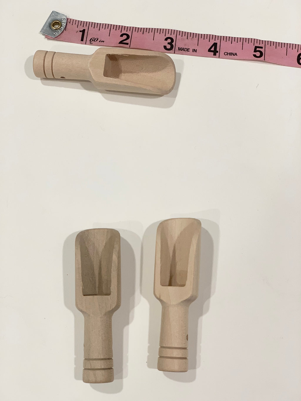 Three natural wooden sensory bin scoops with grooved handles next to a pink measuring tape on white surface