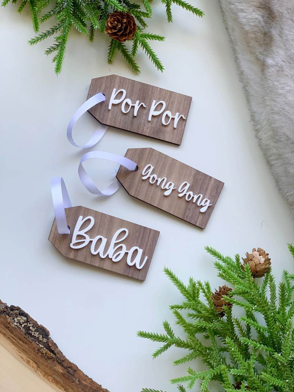 Three custom wooden Christmas gift tags with white cursive names and white ribbons on white surface with pine and fur accents