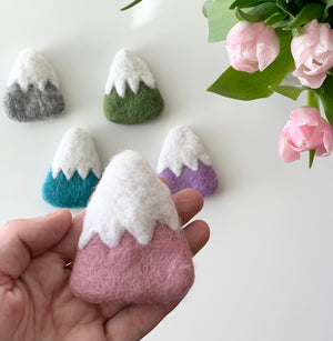 Hand holding one pink felt mountain with white snowcap with four other colorful felt mountains and pink flowers on white surface