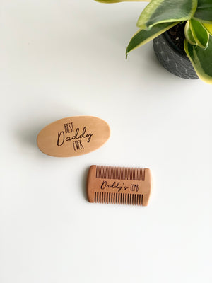 Custom wooden beard brush engraved with Best Daddy Ever and wooden comb engraved Daddy’s Comb on white surface near green plant pot