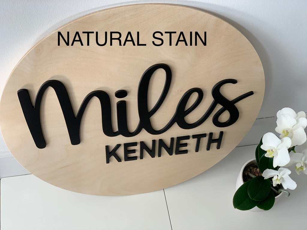 Oval 3D wooden last name sign with natural stain and black cursive and block letters on white surface next to white orchid plant