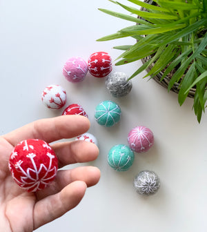 Hand holding red felt snowflake ball with white embroidery among assorted colored felt balls on white surface near green plant
