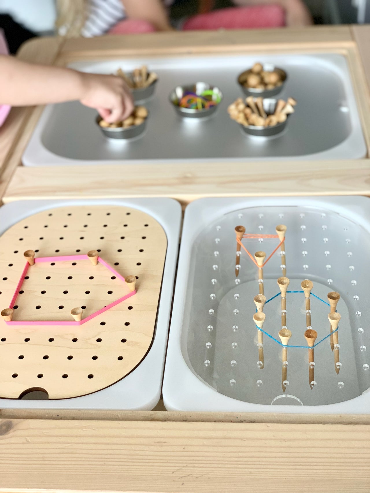 Wooden pegboard and clear board with pegs and colorful rubber bands on wooden table with sensory bins in background