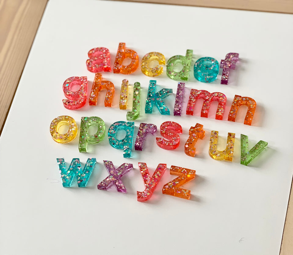 Rainbow glitter resin lowercase alphabet letters arranged in rows on white tray with wooden border