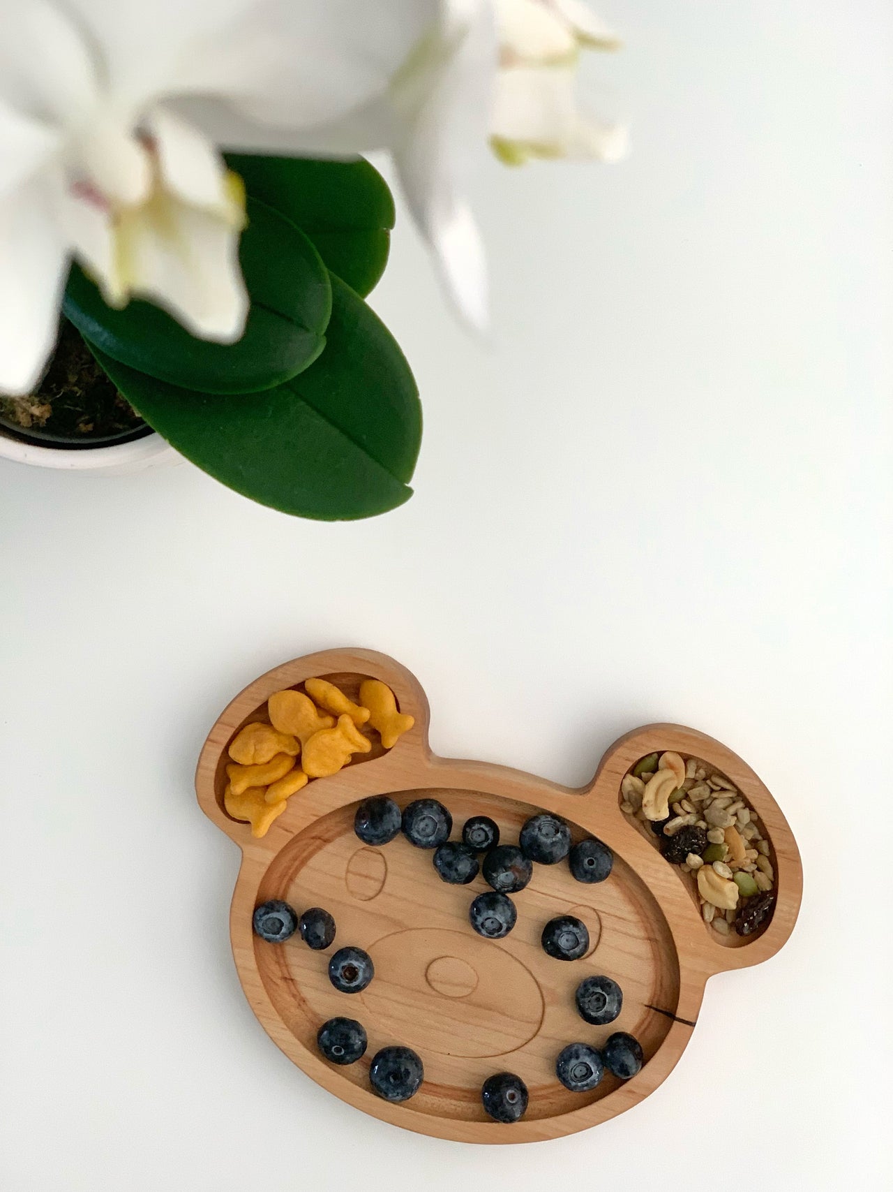 Natural wooden bear-shaped sensory tray with blueberries, goldfish crackers, and mixed nuts on white surface near plant leaves