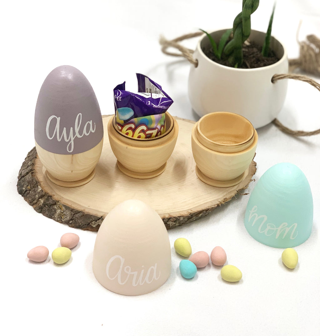 Set of personalized hollow wooden Easter eggs painted in pastel colors with white cursive names on natural wood tray with candy eggs