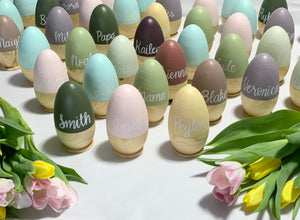 Collection of personalized hollow wooden Easter eggs in pastel colors with white cursive names displayed with tulip flowers