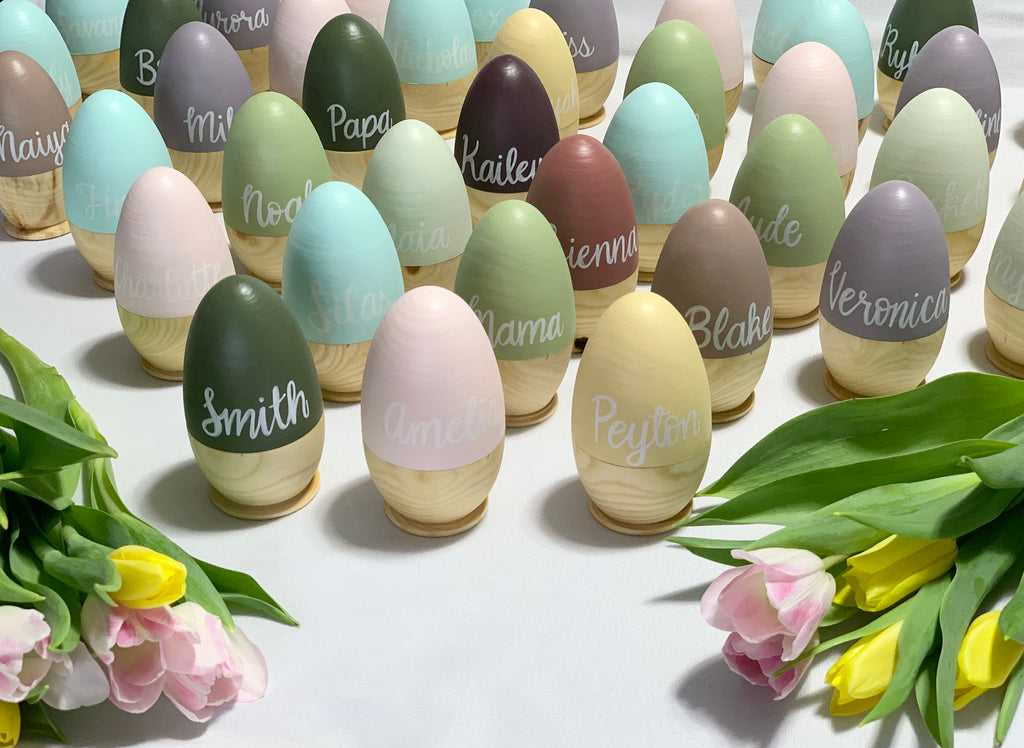 Collection of personalized hollow wooden Easter eggs in pastel colors with white cursive names displayed with tulip flowers
